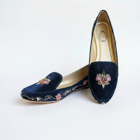 A pair of navy blue velvet slip-on flats with pink floral and gold vine embroidery, displayed on a textured gray surface.
