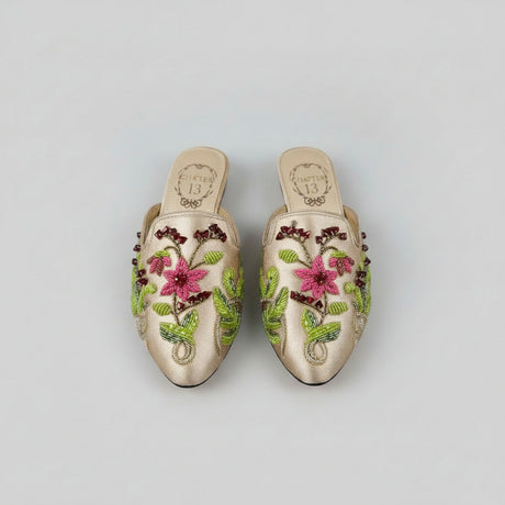 A pair of pointed-toe slip-on mule flats in shimmering champagne satin, decorated with vibrant pink floral embroidery, green leaves, and dark berry accents, resting on a wooden floor.