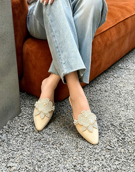 A person seated on an orange-brown sofa wearing pointed-toe slip-on flats with pearl-like floral embellishments on a gray gravel floor.