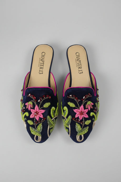 A pair of navy blue velvet slip-on mule flats with colorful floral embroidery in pink, green, and gold, shown on a light wood floor. The brand name 'COSI' is visible on the insoles.
