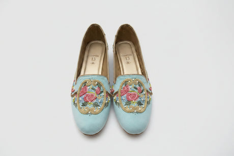 A pair of light blue slip-on shoes with detailed pink floral embroidery and gold braided trim, displayed on a wooden block.