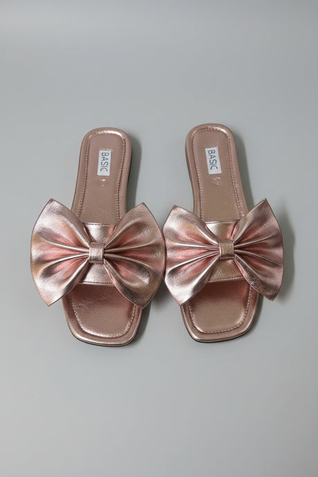 A pair of metallic rose gold slide sandals with large bow details on the toes, displayed on a light surface.